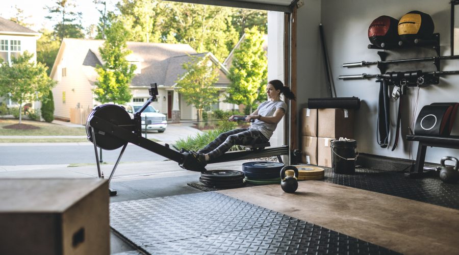Photo of an active woman is exercising on a rowing machine in the home garage gym during covid-19 pandemic. Woman performing powerful pulls and focusing on endurance and cardio improvement.