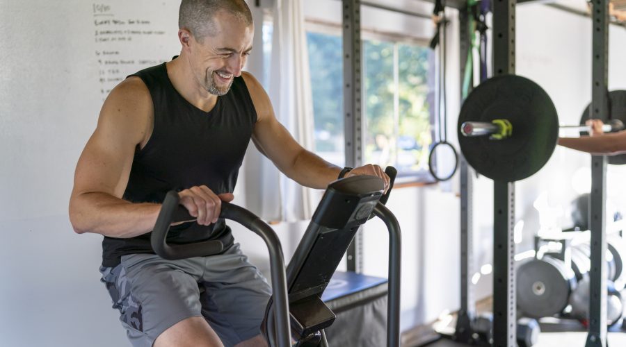 An attractive and fit man smiles while doing a cardio workout on a stationary bike. The man is exercising in a gym he has set up in the garage of his home.