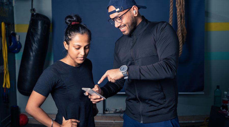 Asian/Indian Woman and personal male trainer making exercise plan on smartphone in gym.